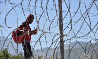 epa09209507 A migrant waits to sneak through the fence in the northern town of Fnideq in an attempt to cross the border from Morocco to the Spanish enclave of Ceuta, in North Africa, 18 May 2021.  At least 5,000 migrants, an unprecedented influx in Spanish authorities said a period of high tension between Madrid and Rabat crept into Ceuta on 17 May, a record for a day. They reached the coast by swimming or walking at low tide from beaches a few kilometers to the south, some using inflatable swimming rings.  EPA/JALAL MORCHIDI