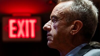 Robert Kennedy Jr. di profilo con segnale EXIT rosso luminoso sullo sfondo - Analisi critica The Lancet sulla Sanità USA