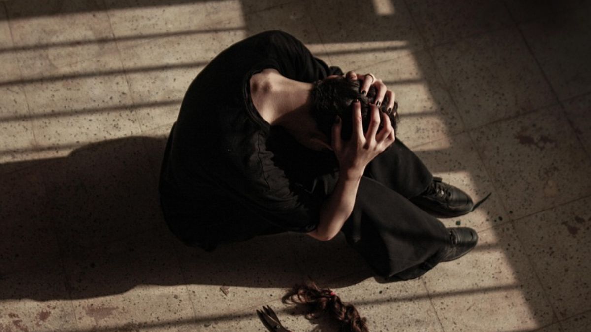 Donna in abiti neri seduta a terra con la testa tra le mani, capelli tagliati sul pavimento, in un ambiente con luce filtrata